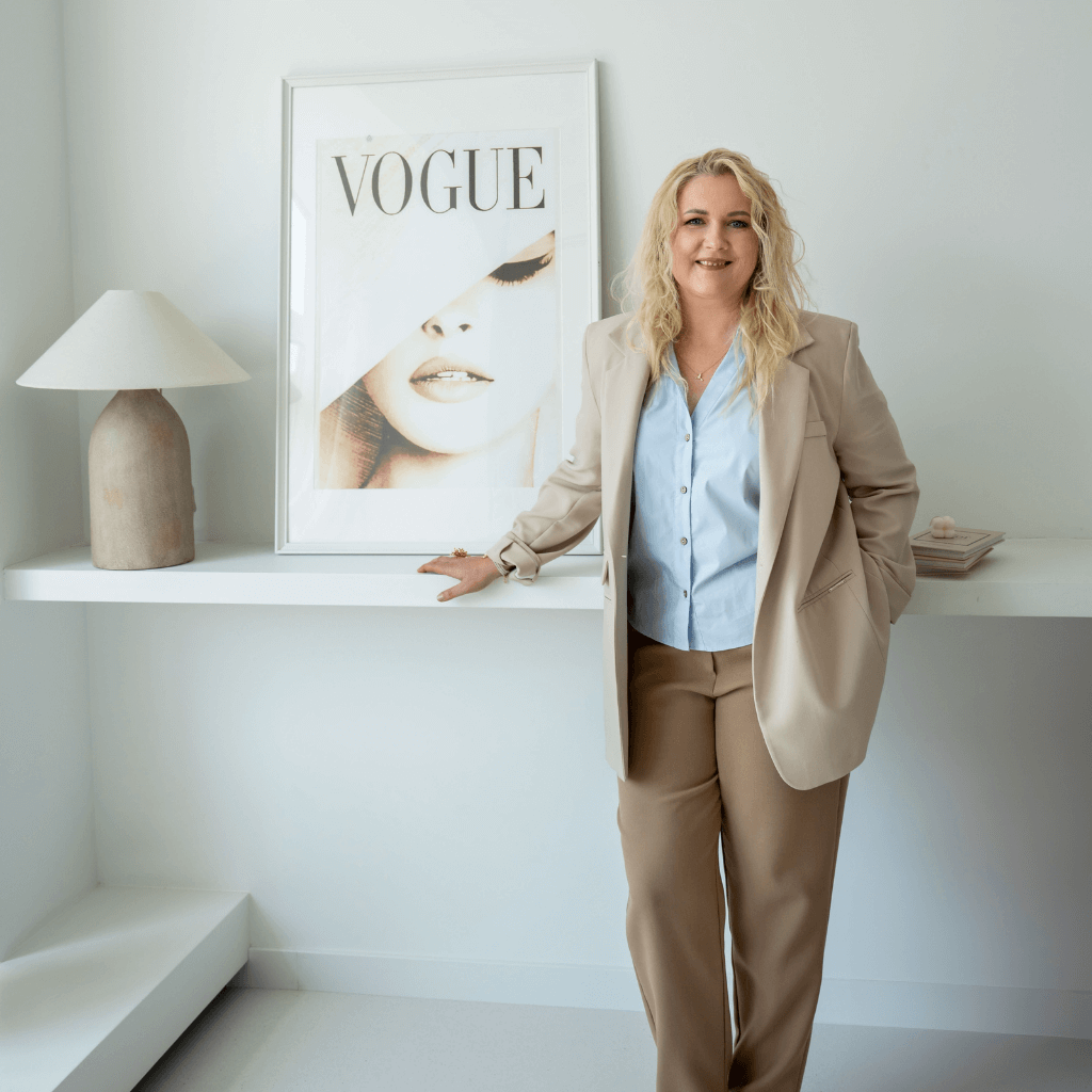 Hipnoterapeutė stovinti šviesioje, minimalistinėje erdvėje šalia „Vogue“ plakato – šiltas ir profesionalus portretas