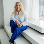 Simona Roganova sėdi ant baltų laiptų prie lango ir šypsosi, vilkėdama baltus marškinius ir mėlynas kelnes