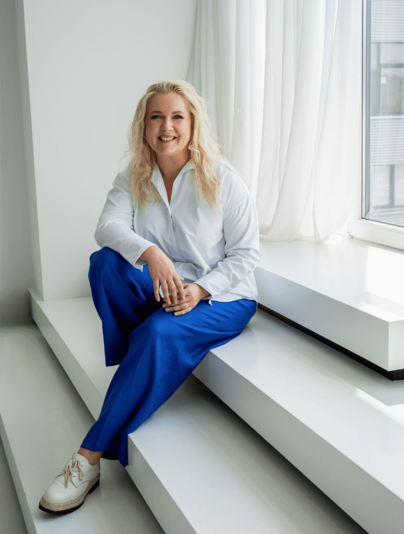 Simona Roganova sėdi ant baltų laiptų prie lango ir šypsosi, vilkėdama baltus marškinius ir mėlynas kelnes