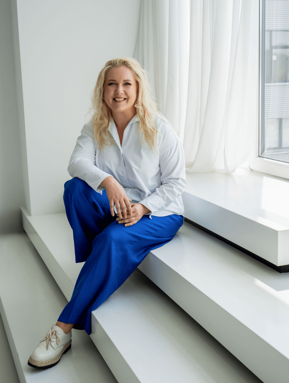 Simona Roganova sėdi ant baltų laiptų prie lango ir šypsosi, vilkėdama baltus marškinius ir mėlynas kelnes